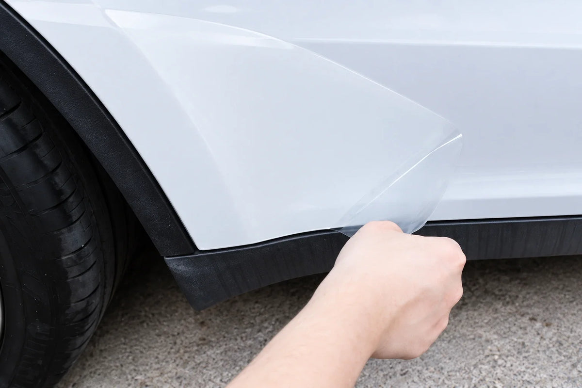 AI-assisted close-up based on a clear paint protection film being applied to a white Tesla lower-door area, used as an editorial illustration of a DIY prep workflow.